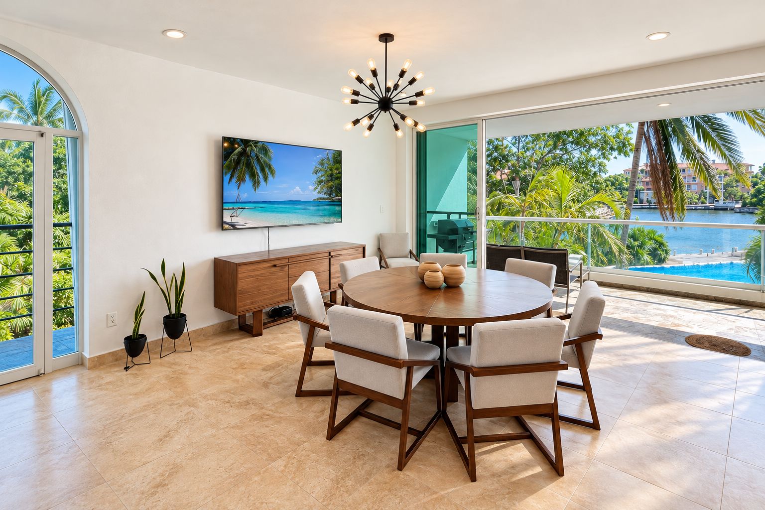A spacious dining area with a round table, chairs, and a view of a pool and palm trees.