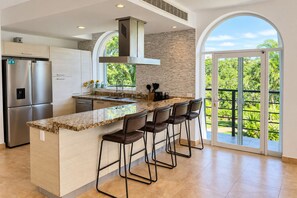 A modern kitchen with a central island, stainless steel appliances, and a large window overlooking greenery. - High-End Waterfront Condo Secrets Waters (Puerto Aventuras)