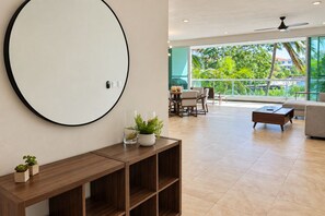 A modern living room with a large round mirror, a wooden console table with potted plants, a dining area with chairs and a table, and a view of greenery through large windows. - High-End Waterfront Condo Secrets Waters (Puerto Aventuras)