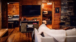 A cozy cabin interior with a wooden interior, a flat-screen TV, a fireplace, and a bed with a white blanket.