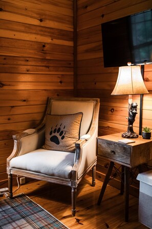 Une pièce aux murs lambrés, avec un fauteuil moelleux agrémenté d’un coussin à motif d’empreinte de patte, une petite table sur laquelle se trouve une lampe et une statue décorative, et un téléviseur fixé au mur.