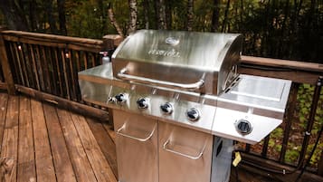 A stainless steel NEXGRILL barbecue grill with four burners, a side burner, and a built-in thermometer, set on a wooden deck with a metal railing.