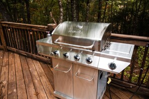 Un barbecue NEXGRILL en acier inoxydable, doté de quatre brûleurs, d’un brûleur latéral et d’un thermomètre intégré, installé sur une terrasse en bois avec une rambarde en métal.