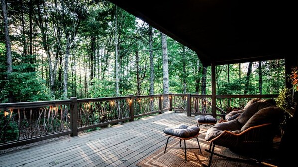 A wooden deck with a railing, two cushioned chairs, and a view of a forest.