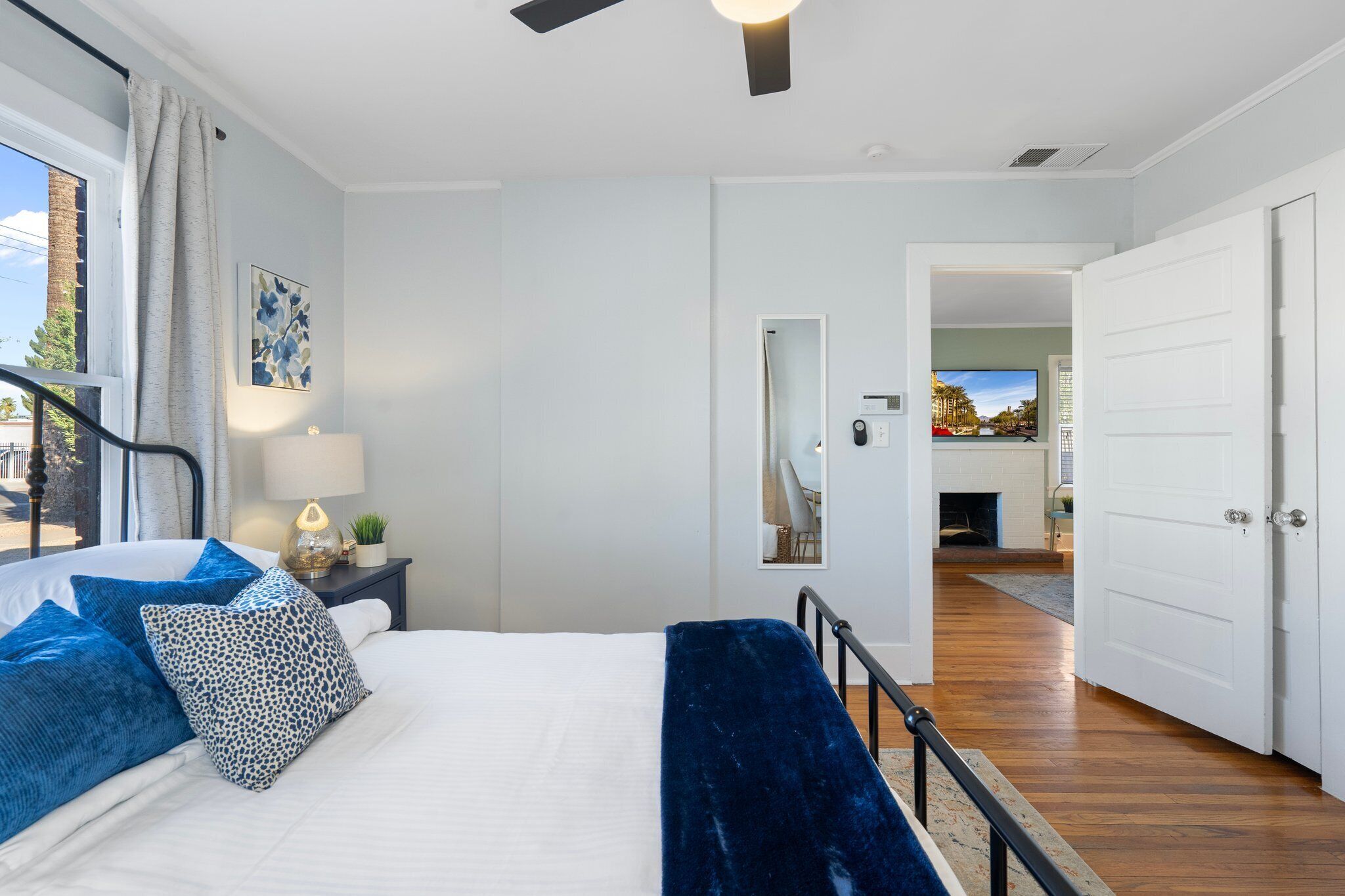 Quarto com cama, mesa de cabeceira, luminária, janela com cortinas, ventilador de teto e vista para uma sala de estar com lareira.