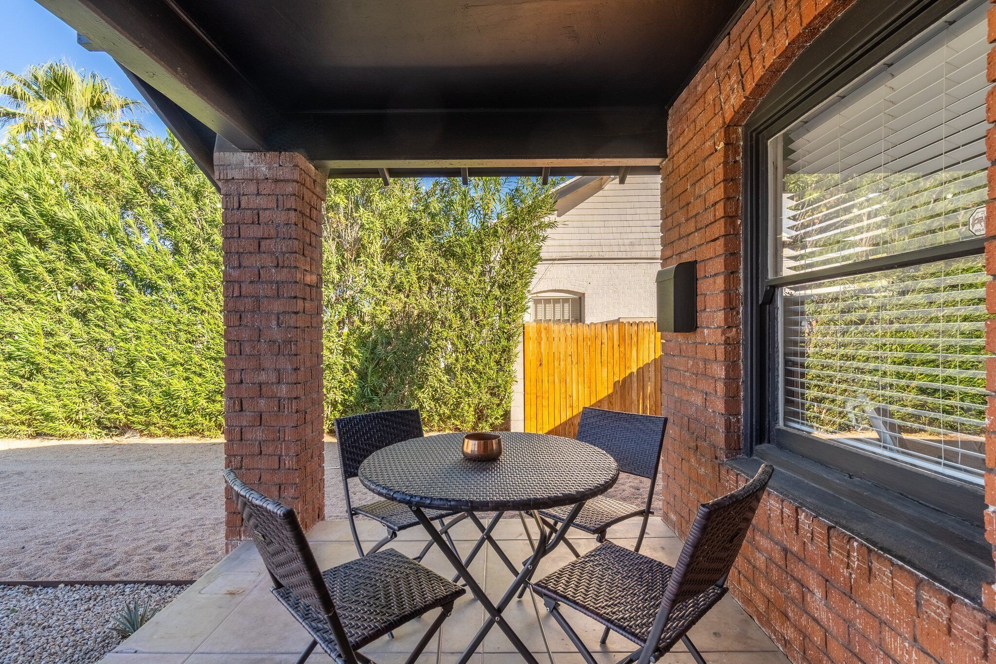 An outdoor patio with a round table and four chairs, a brick pillar, and a window with blinds.