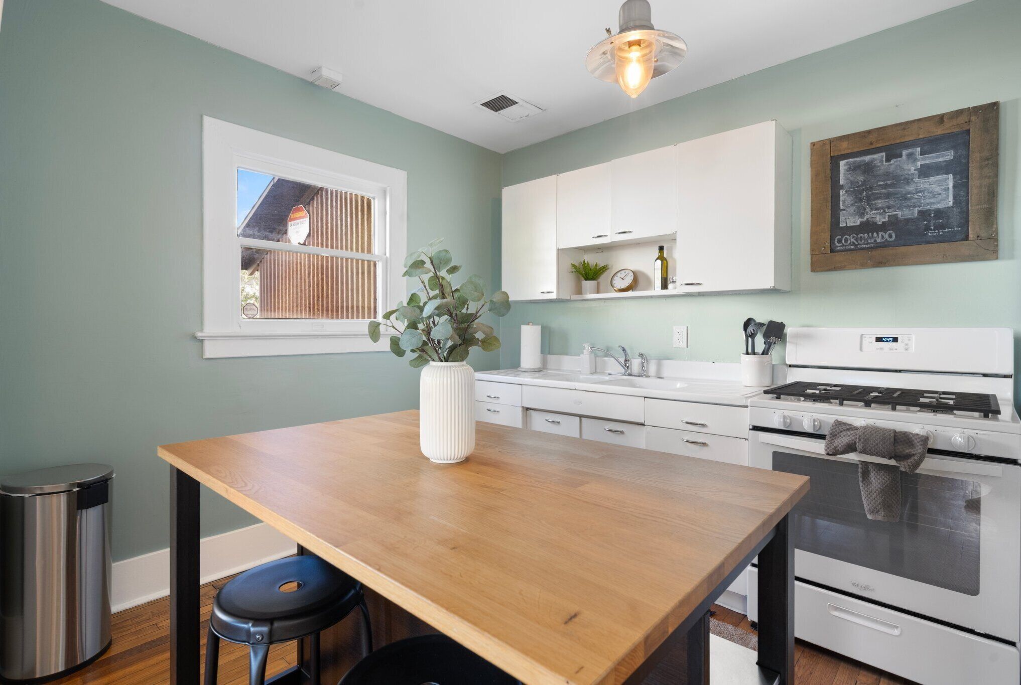 A kitchen with a wooden table, white cabinets, a stove, and a window with blinds.