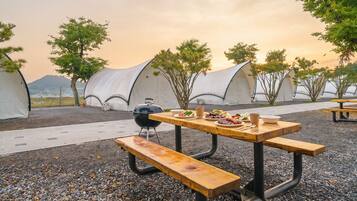 A picnic setup with a table, benches, and a grill, surrounded by tents and trees.