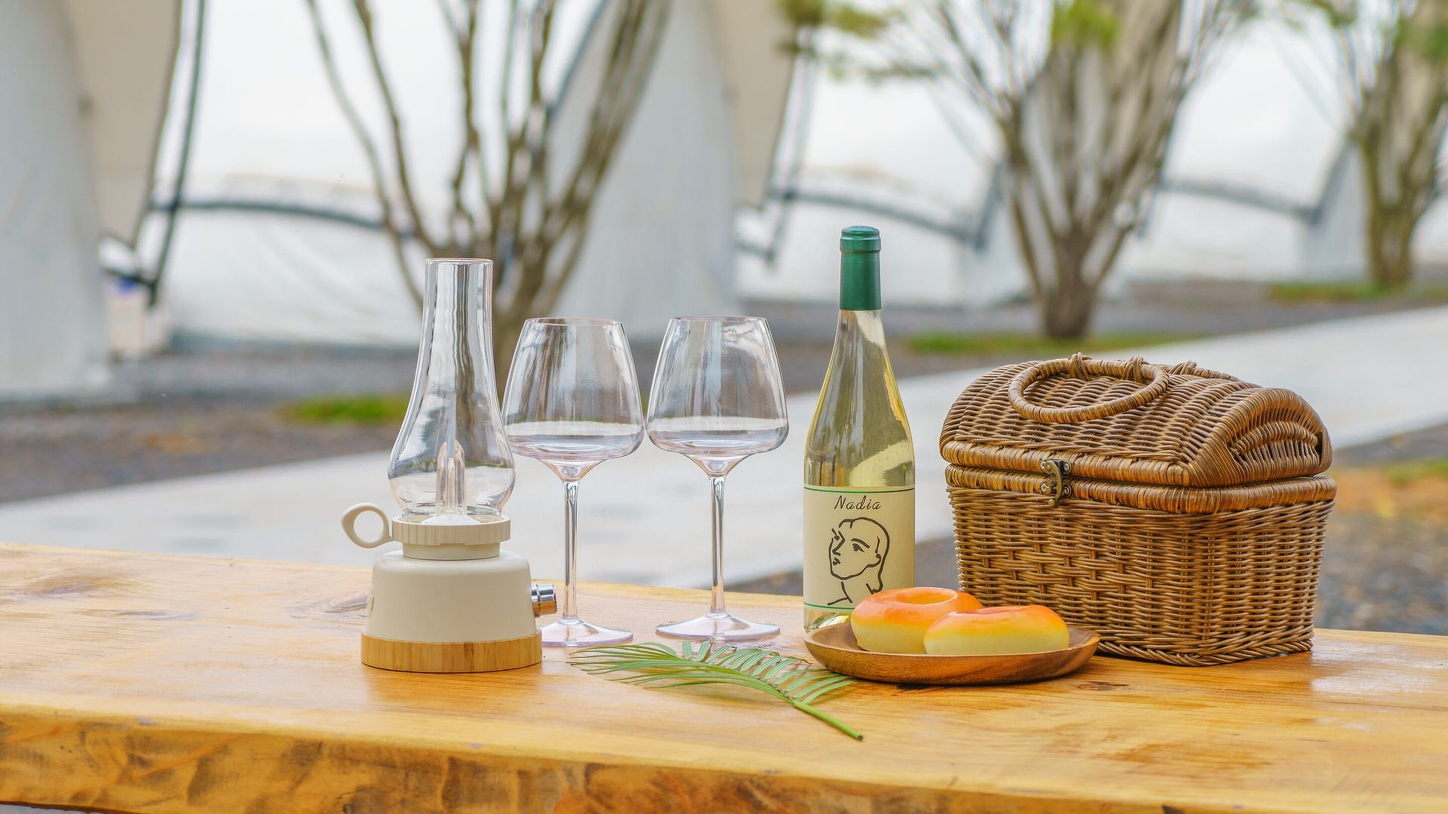 A wooden table with a bottle of wine, three wine glasses, a wicker basket, and a small plate with pastries.
