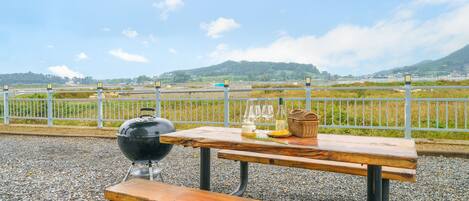 An outdoor picnic setup with a grill, picnic table, and bottles of wine, overlooking a scenic landscape with hills and a fence.