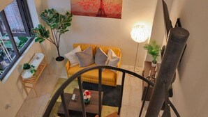 Una sala de estar con un sofá amarillo, una escalera, una planta en maceta y un cuadro de la Torre Eiffel.