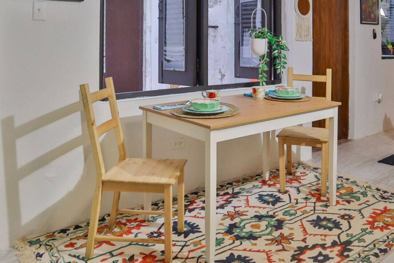 A dining area with a wooden table and chairs, a colorful patterned rug, and a window with shutters.