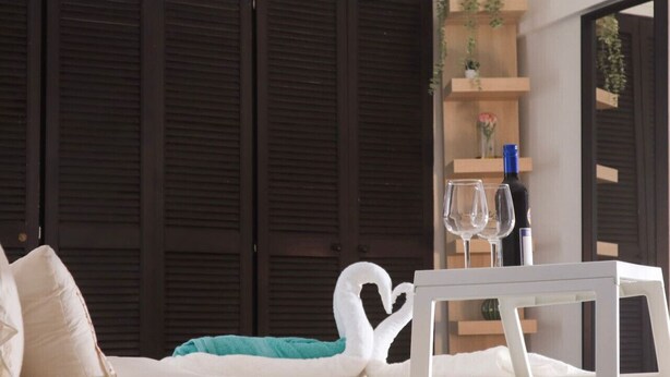 Un dormitorio con cama, almohadas blancas, una mesita de noche blanca con vaso y botella, una toalla con forma de corazón y contraventanas de madera oscura.