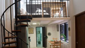 Un apartamento de dos pisos con una escalera espiral, un balcón con una silla blanca, una zona de comedor con una mesa y sillas, y una ventana con una planta.
