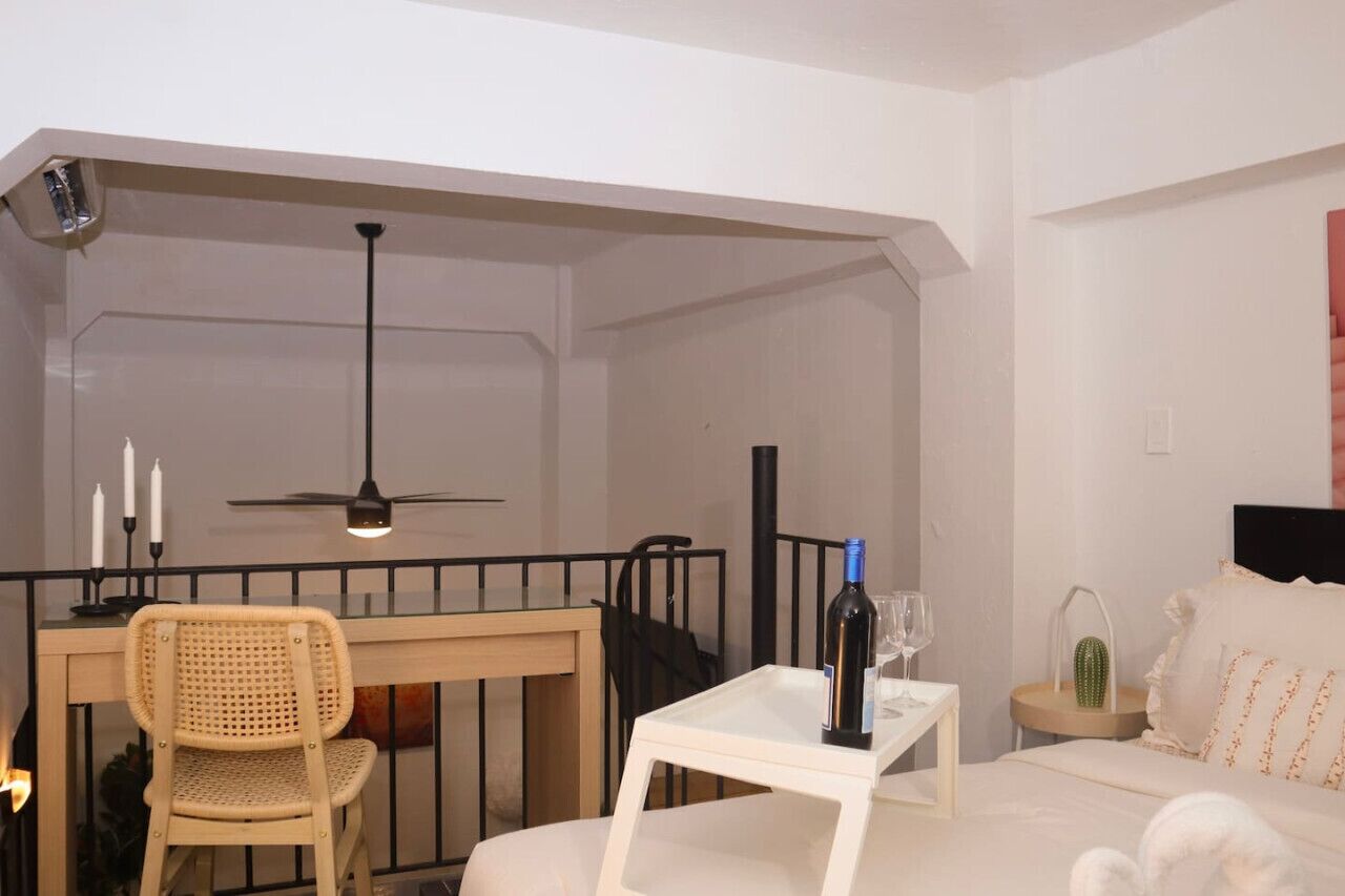 A bedroom with a bed, a bedside table with a wine bottle, a chair, a desk, and a ceiling fan.