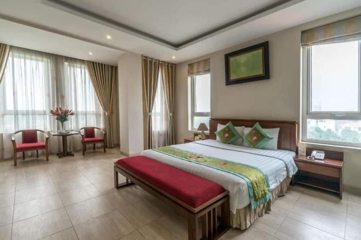 Une chambre d’hôtel avec un grand lit, une banquette, deux chaises, une petite table et une fenêtre avec des rideaux.