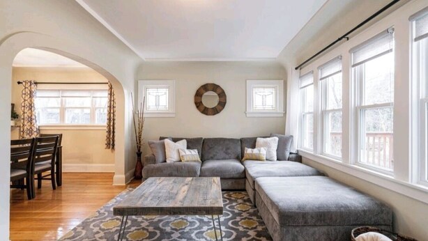 A living room with a grey sectional sofa, a patterned rug, and a wooden coffee table. There are two windows with white frames and a ceiling light.