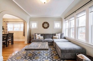 Una sala de estar con un sofá seccional gris, un tapete estampado y una mesa de centro de madera. Hay dos ventanas con marcos blancos y una lámpara de techo.