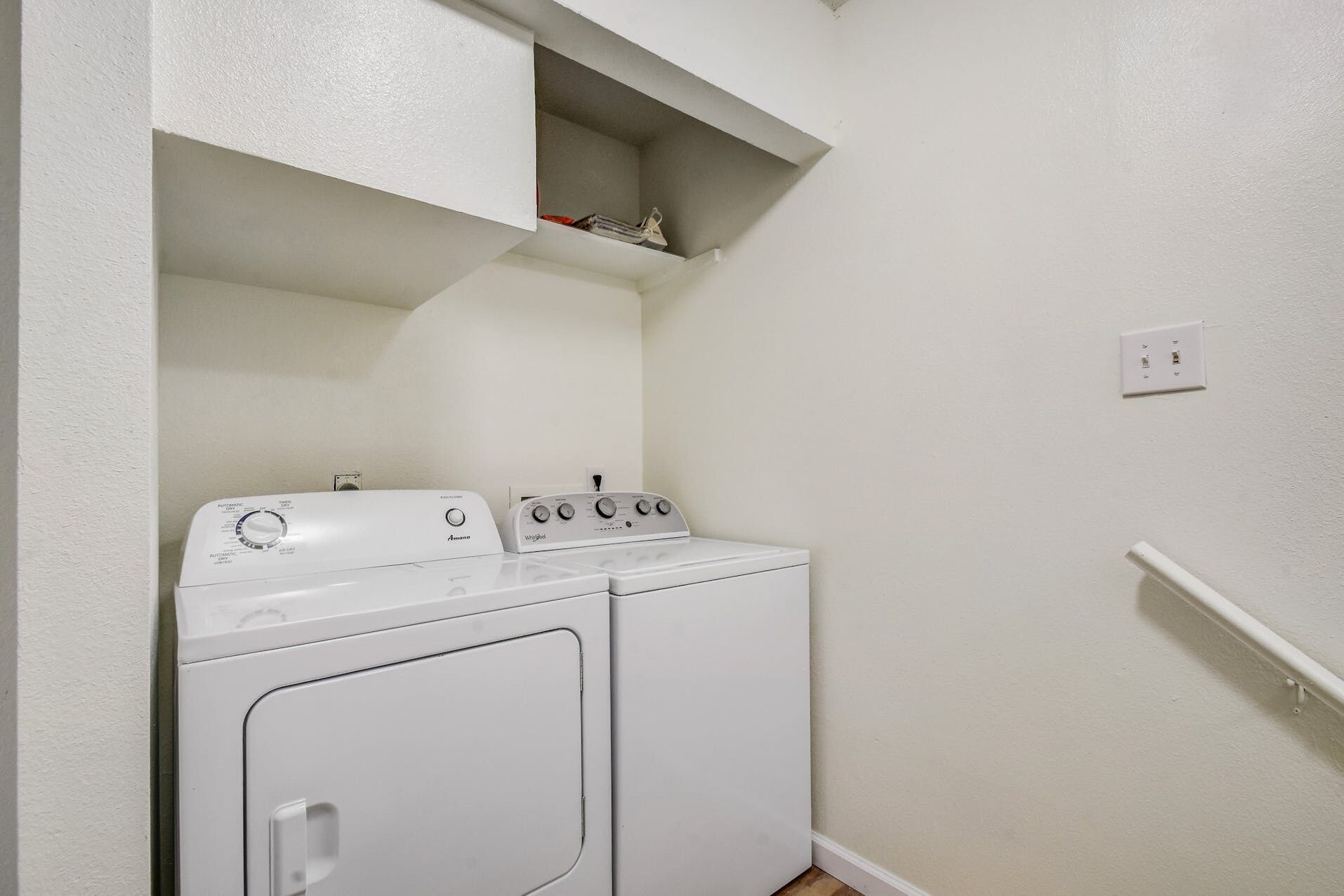 A laundry room with a washer and dryer, a shelf above the dryer, and a power outlet on the wall.