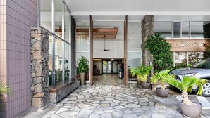 L’entrée d’un hôtel, avec un chemin bordé de dalles de pierre, de grandes fenêtres et des plantes en pot.