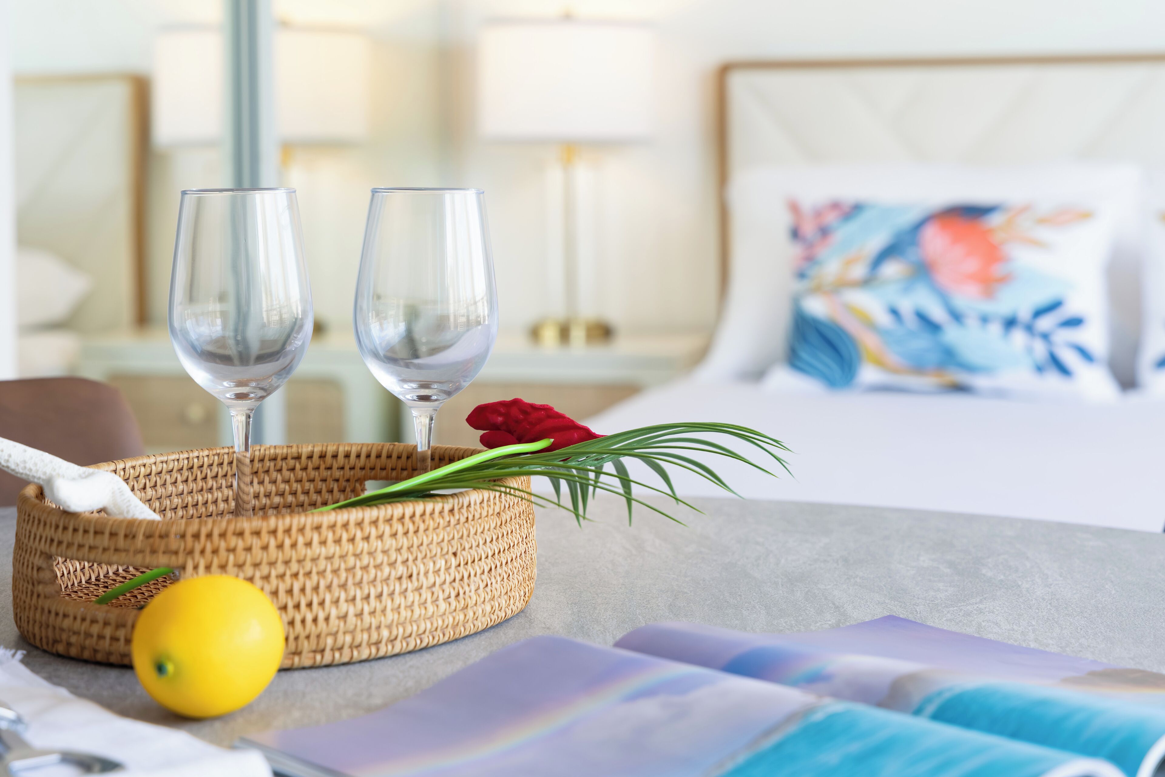 Une table soigneusement dressée, avec deux verres à vin, un citron et un panier tressé contenant une rose et des feuilles vertes.