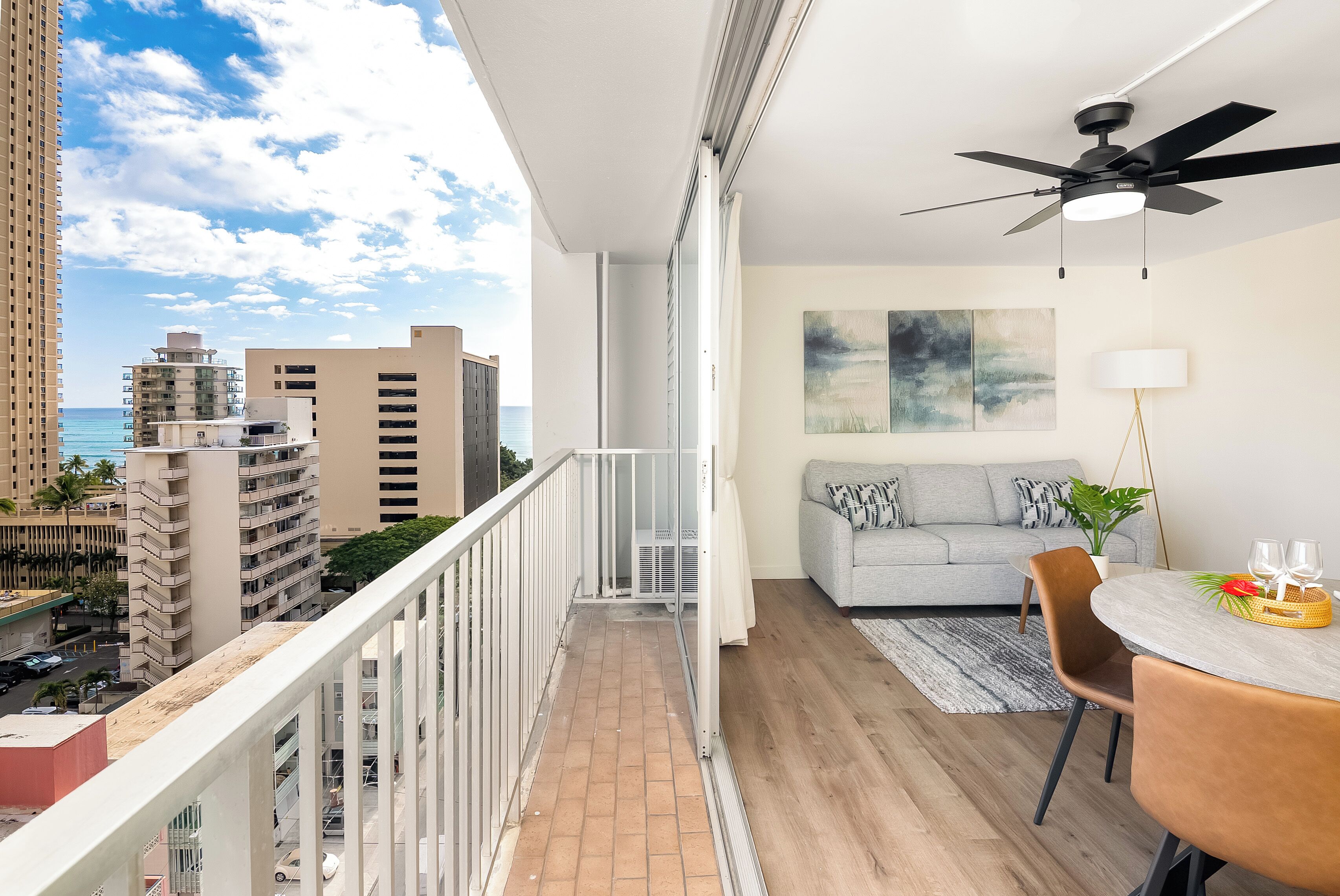 A balcony with a city view, a grey sofa, a dining table with chairs, and a ceiling fan.
