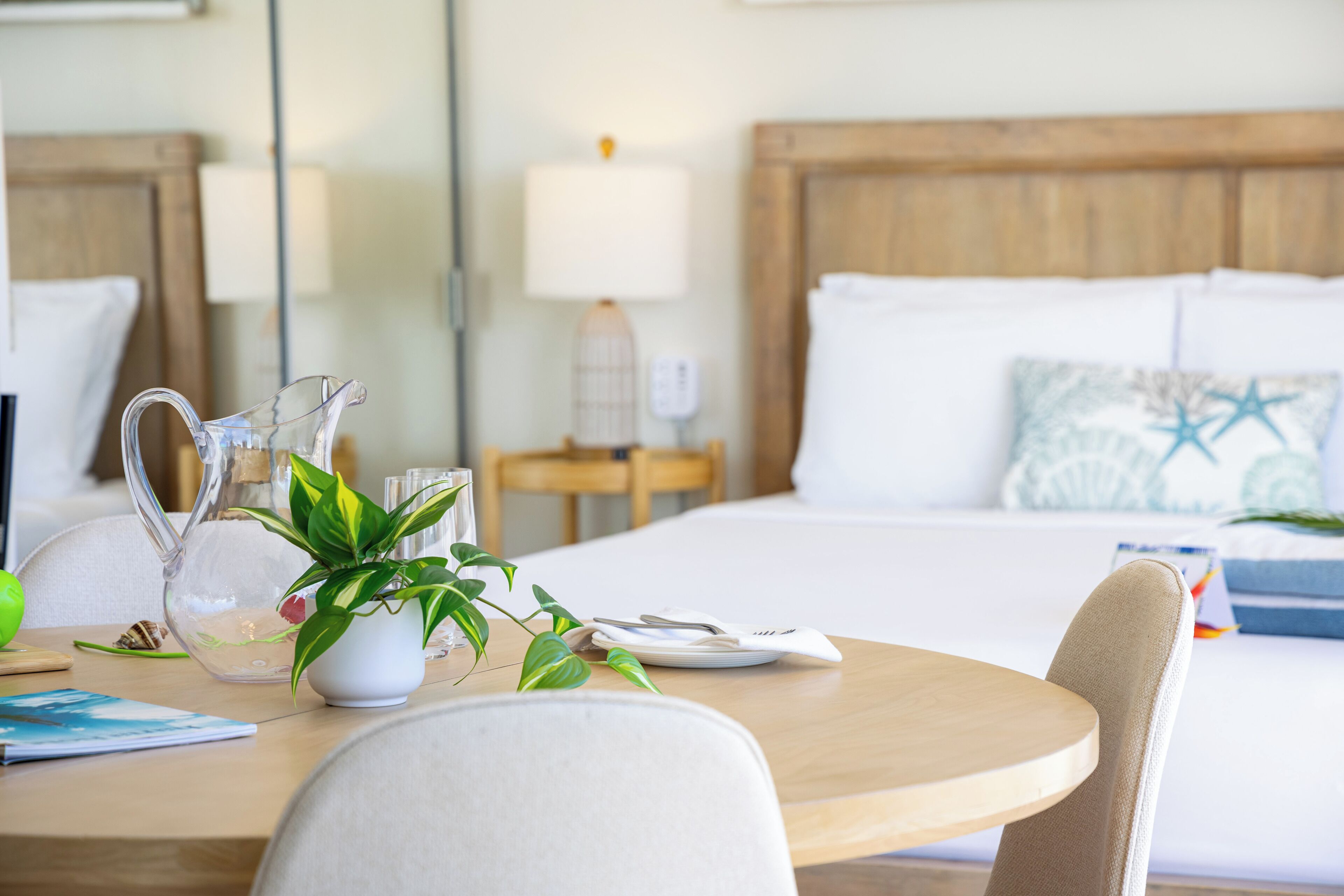 A hotel room with a wooden headboard, a bed with white linens, a bedside table with a lamp, a pitcher, and a potted plant.