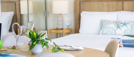 A hotel room with a wooden headboard, a bed with white linens, a bedside table with a lamp, a pitcher, and a potted plant.