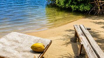 Ein Strand mit einer Holzbank und einer Liege, freier Blick auf das Wasser und ein Haus im Hintergrund.