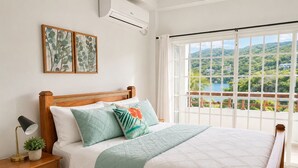 Ein Schlafzimmer mit einem Bett, einem Nachttisch, einer Lampe und Blick auf eine üppige Landschaft durch große Fenster.