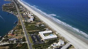 An aerial view of a coastal area with a long stretch of beach, a road parallel to the shore, and several buildings including a large modern structure.