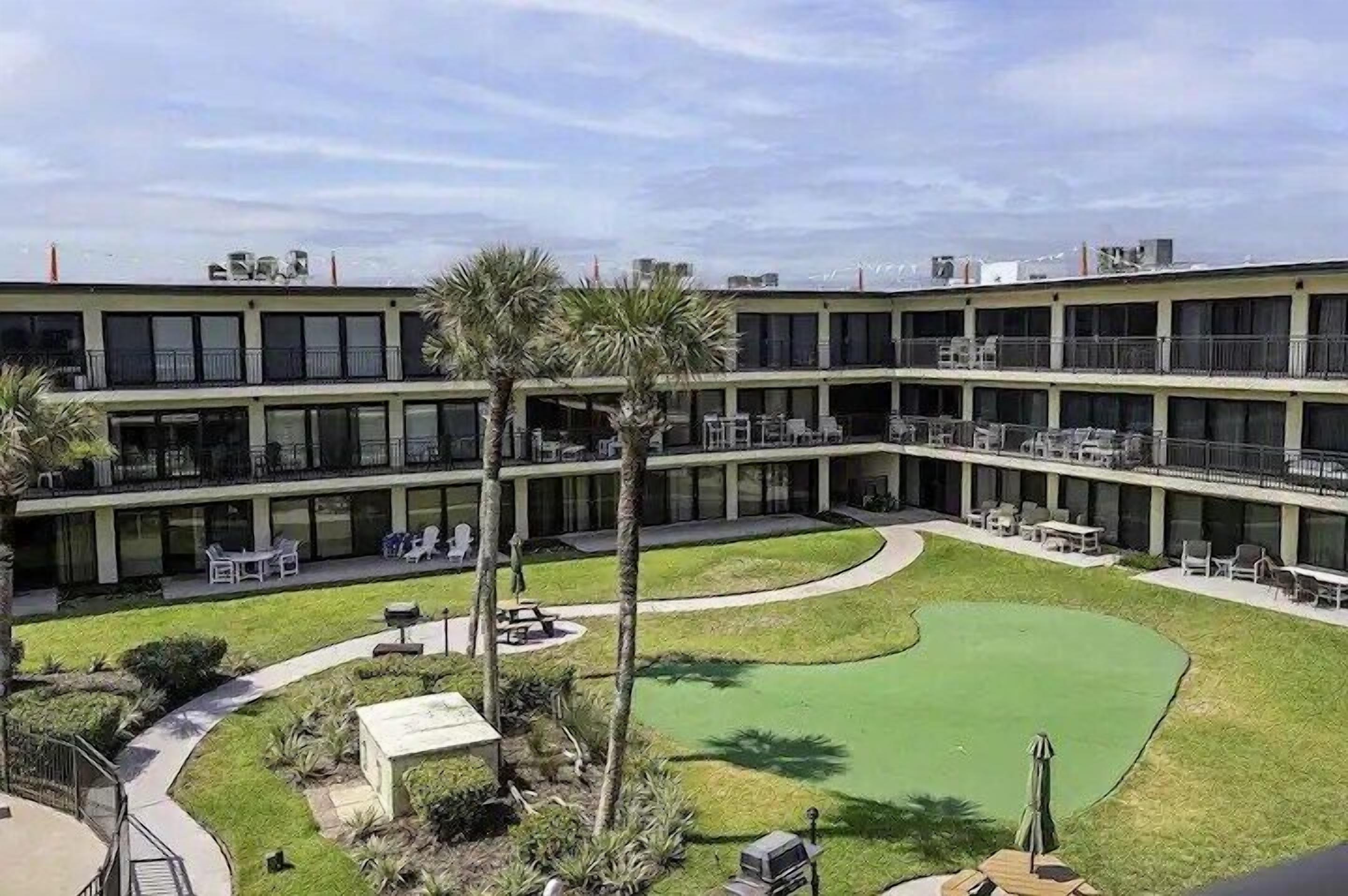 A multi-story hotel with a courtyard featuring a green lawn, outdoor seating, and palm trees.