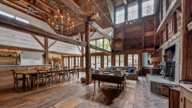 A rustic interior with wooden floors, a dining area with wooden tables and chairs, a fireplace, and large windows allowing natural light. - Renovated Grand Woodstock Barn (Bearsville)