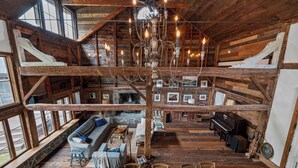 A rustic cabin with a stone fireplace, wooden beams, a chandelier, a piano, and a cozy seating area with a sofa and chairs. - Renovated Grand Woodstock Barn (Bearsville)