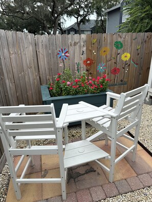A patio with a white table and chairs, a wooden fence with painted flowers, and a planter with red flowers.