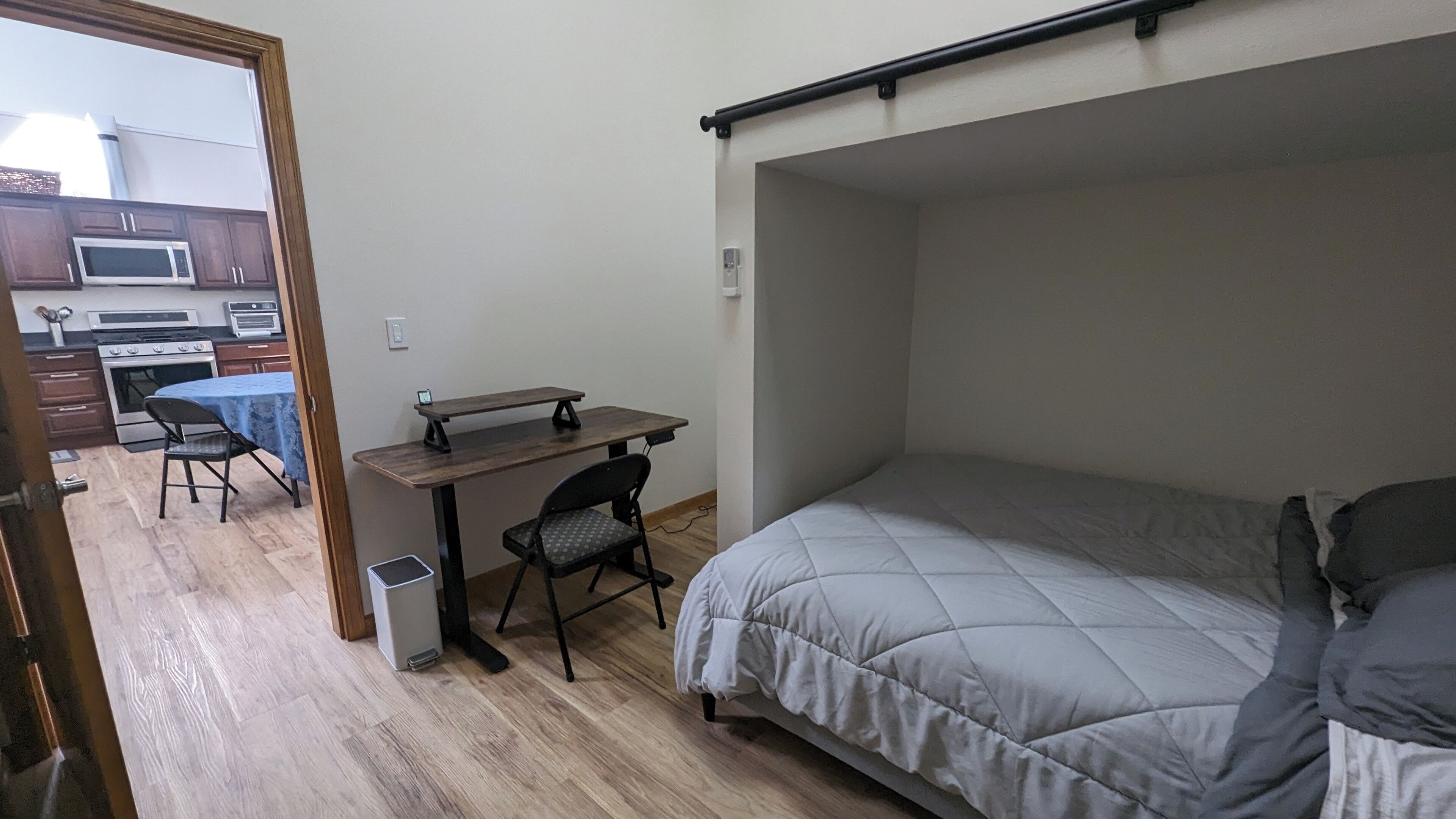 A compact bedroom with a bed, a desk, and a chair. A kitchenette with a microwave and stove is visible in the adjacent room.