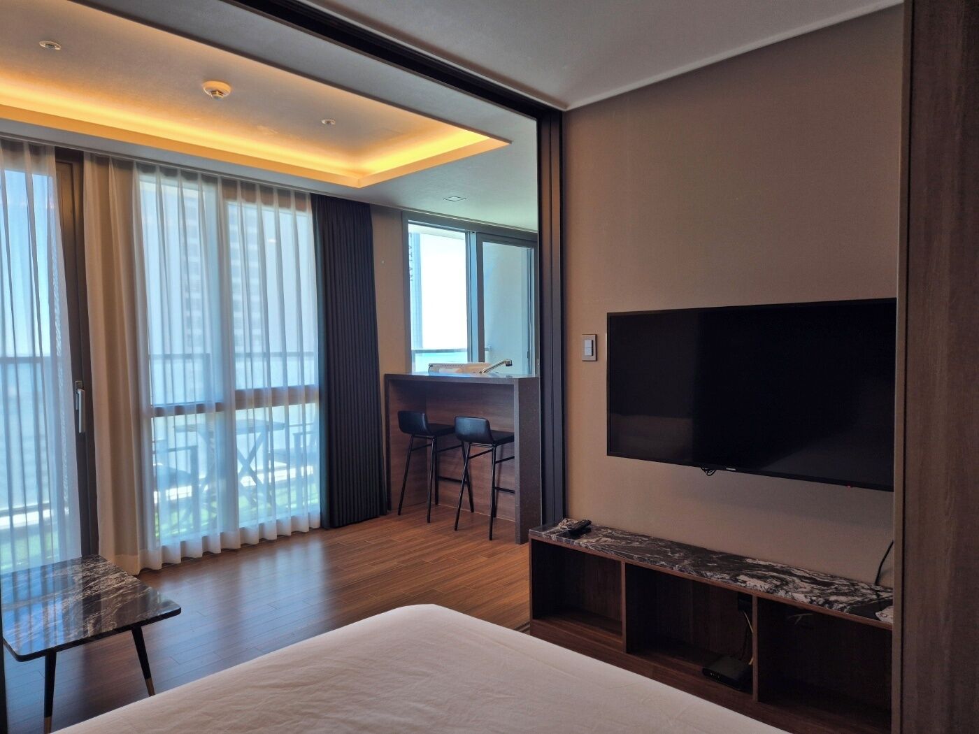 Una habitación de hotel moderna con una cama grande, un televisor de pantalla plana montado en la pared, un escritorio pequeño con dos sillas y un balcón con cortinas.