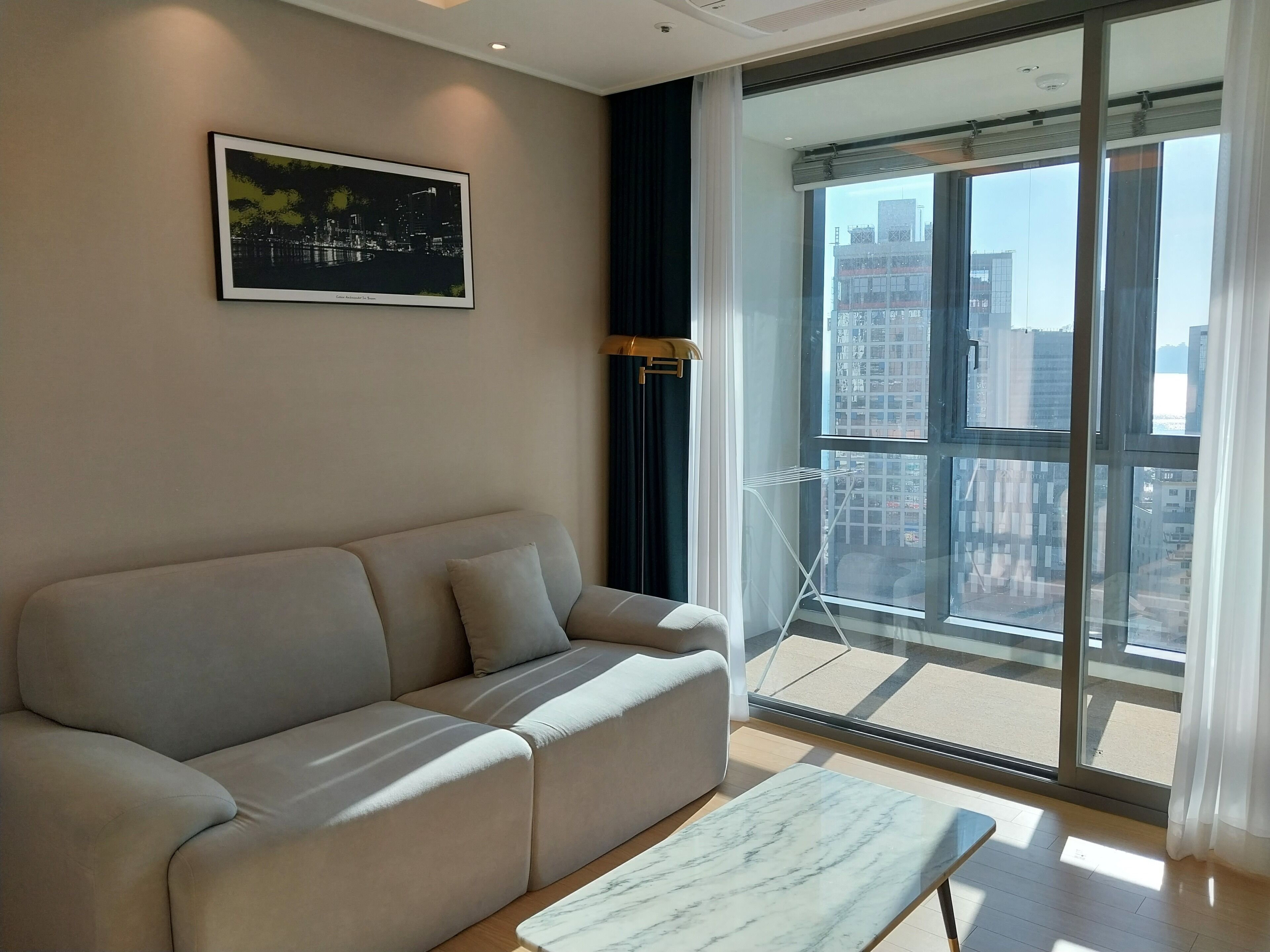 A modern living room with a beige sofa, a marble coffee table, and a large window offering a city view.