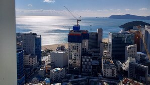 A high-rise building with a construction crane, a beach, and a mountainous landscape in the background. - The solid residence - Elbon by haeundae (Busan)