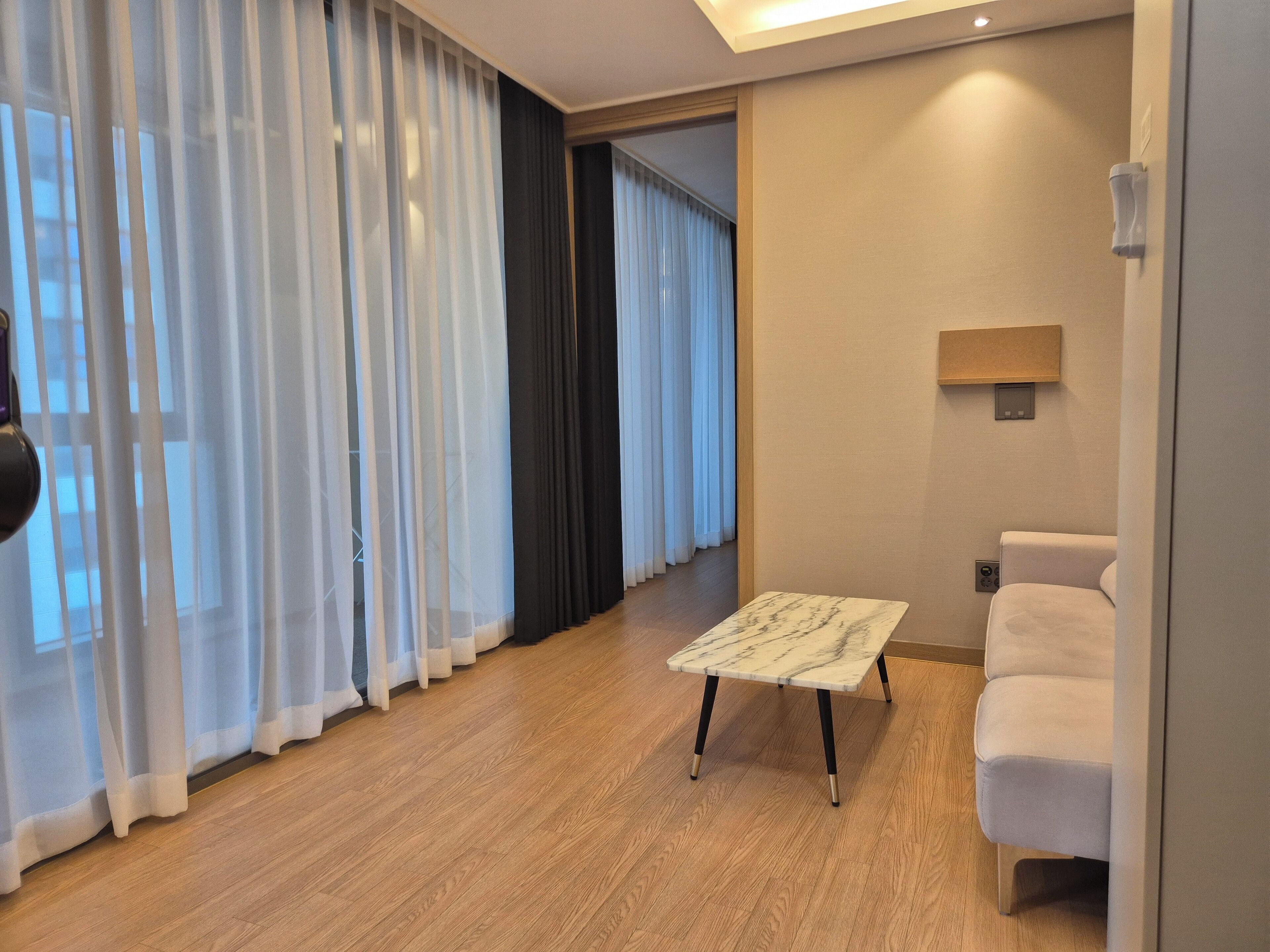 A modern hotel room with a wooden floor, a marble coffee table, a white sofa, and a large window with sheer curtains.