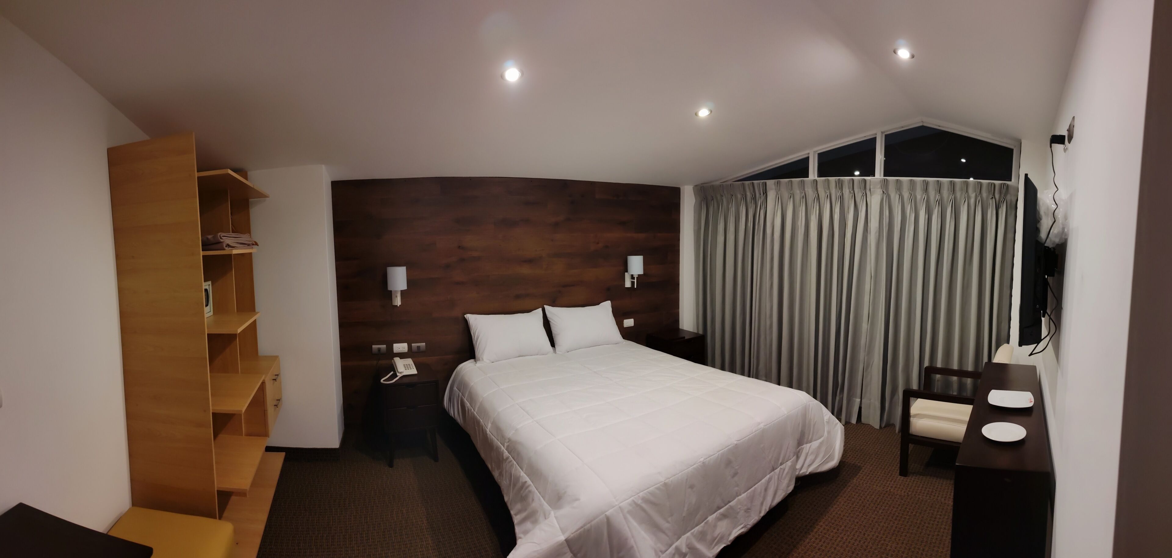 A hotel room with a large bed, a wooden shelf unit, a desk with a chair, and a window with curtains.