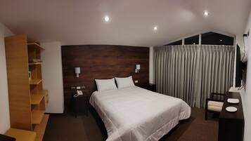 A hotel room with a large bed, a wooden shelf unit, a desk with a chair, and a window with curtains.