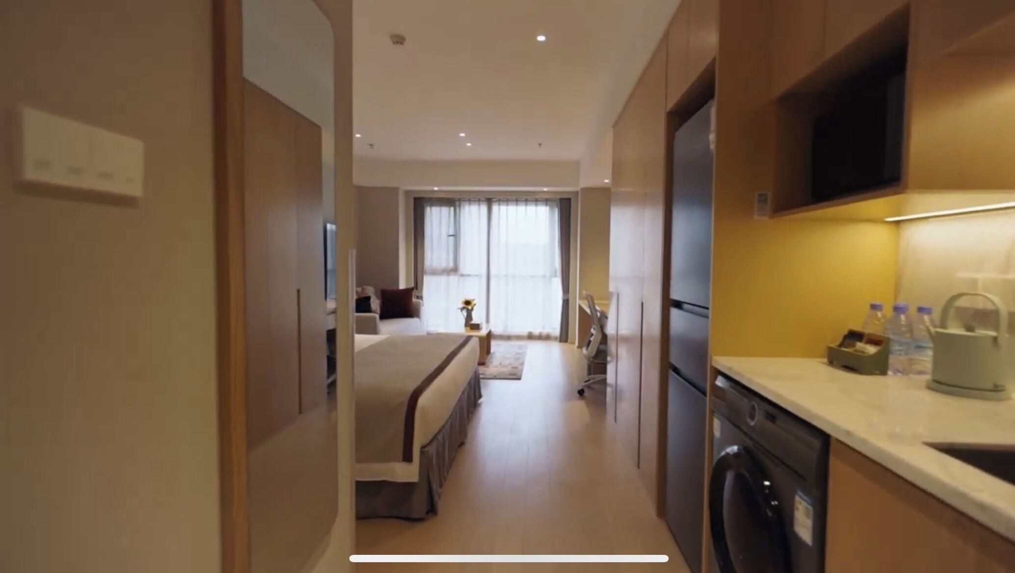 A hotel room with a bed, a desk, a chair, a washing machine, and a kitchen area with a microwave and a kettle.