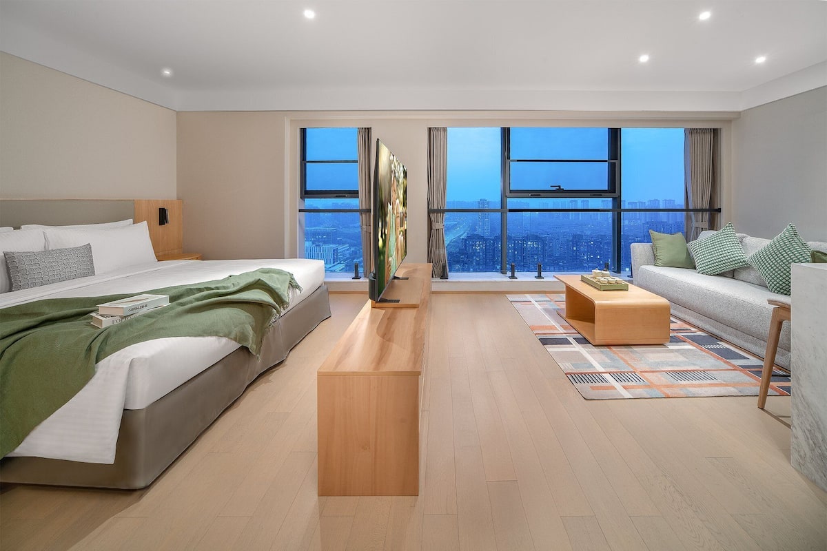 Una habitación de hotel moderna con una cama grande, un sofá, una mesa de centro y vistas de la ciudad a través de amplias ventanas.