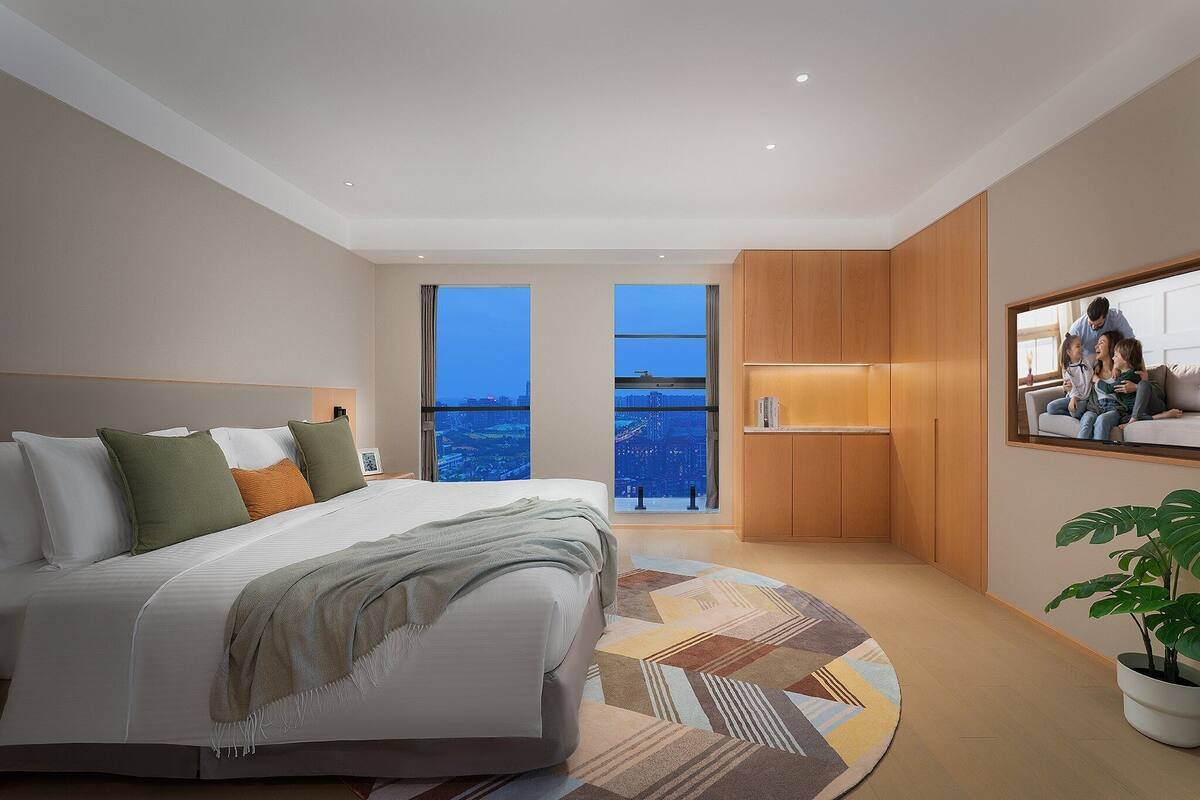Un dormitorio moderno con una cama grande, vistas de la ciudad a través de las ventanas y un pequeño rincón de descanso con una planta.