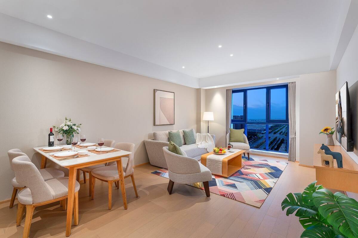 Una sala de estar moderna con comedor, un conjunto de sofás, una mesa de centro y un ventanal con vistas a la ciudad.