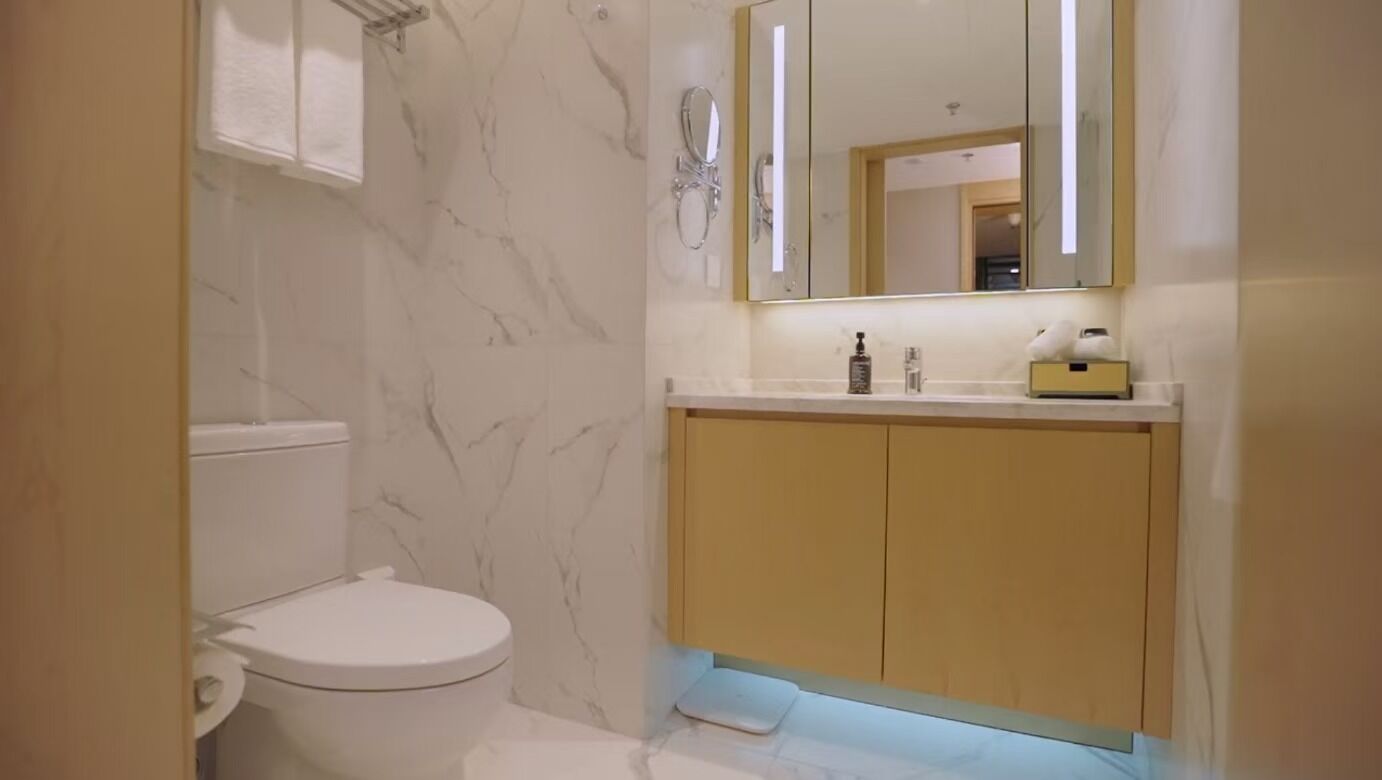A bathroom with a toilet, marble walls, a sink with a mirror, and a cabinet.