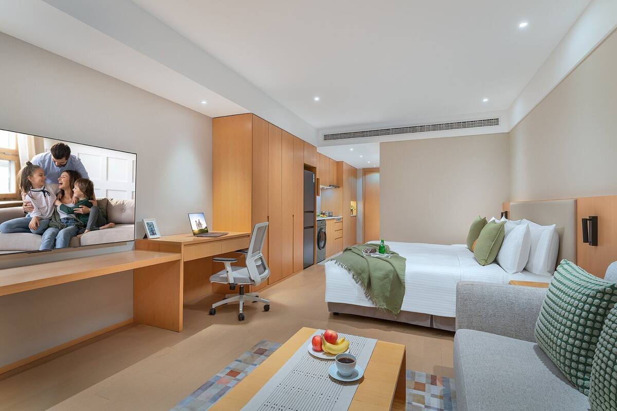 Una habitación de hotel moderna con cama, escritorio, silla, televisión y un rincón de comedor con frutas y una taza.