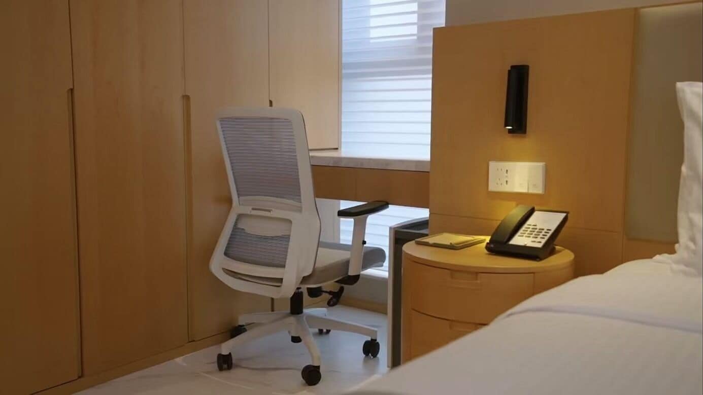 Une chambre d’hôtel avec une chaise de bureau blanche, une table de chevet avec un téléphone et un lit avec du linge de lit blanc.