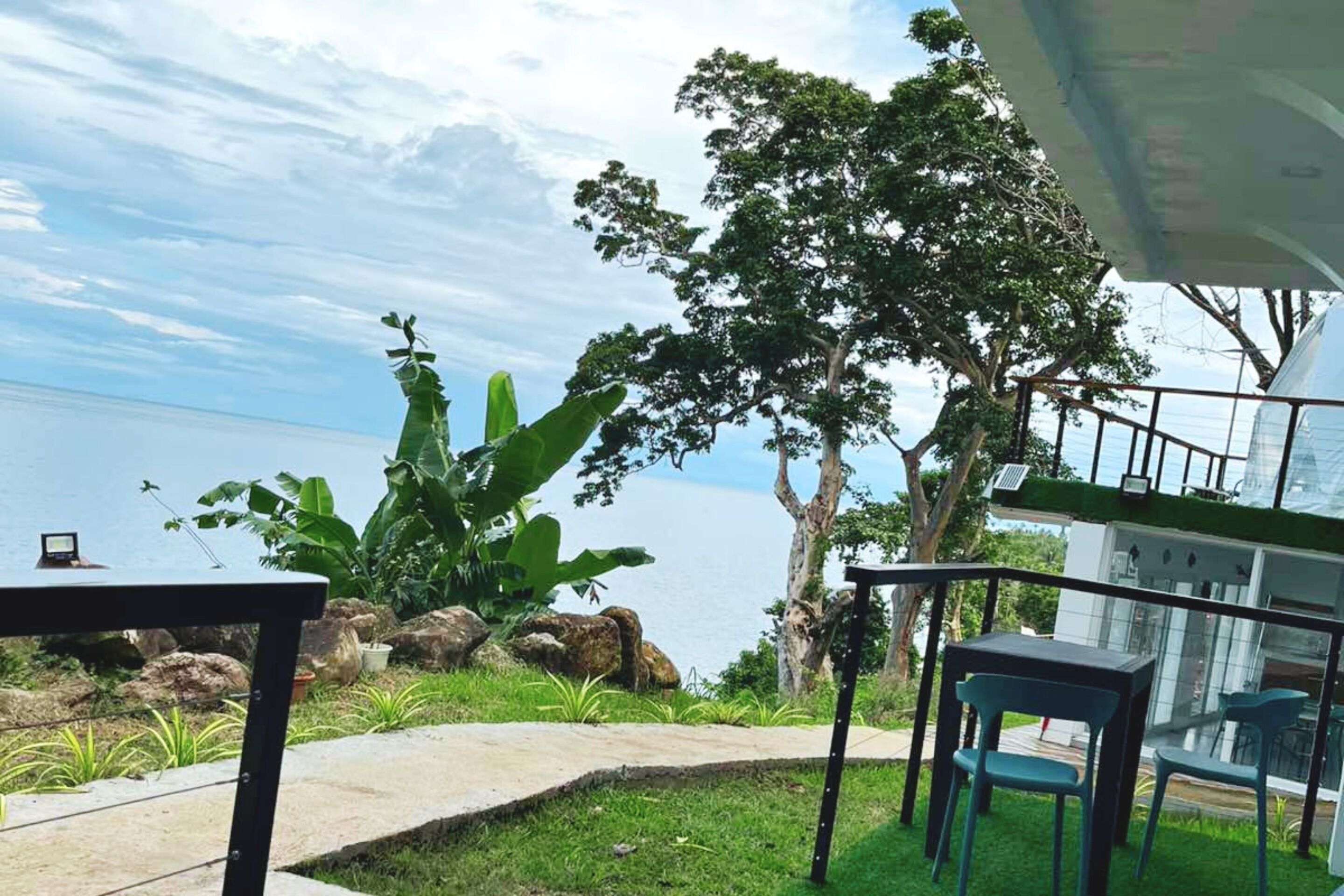 Una terraza con vista al mar, que incluye una mesa y sillas, rodeada de follaje y rocas.